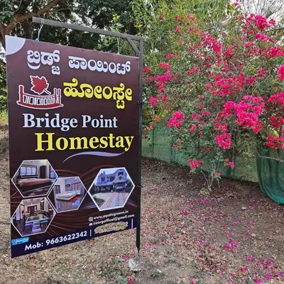 a sign for a bridge point homesteadery next to flowers at MysticGreens Guest House! in Kushālnagar
