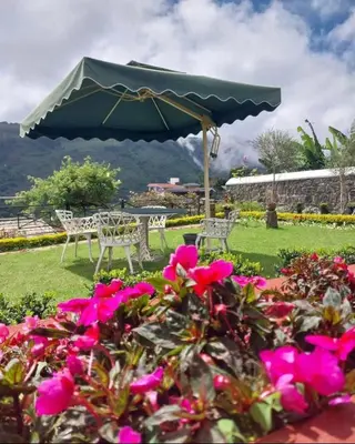 a table and chairs with a green umbrella and pink flowers at Ivy Cottage Kodaikanal in Kodaikānāl